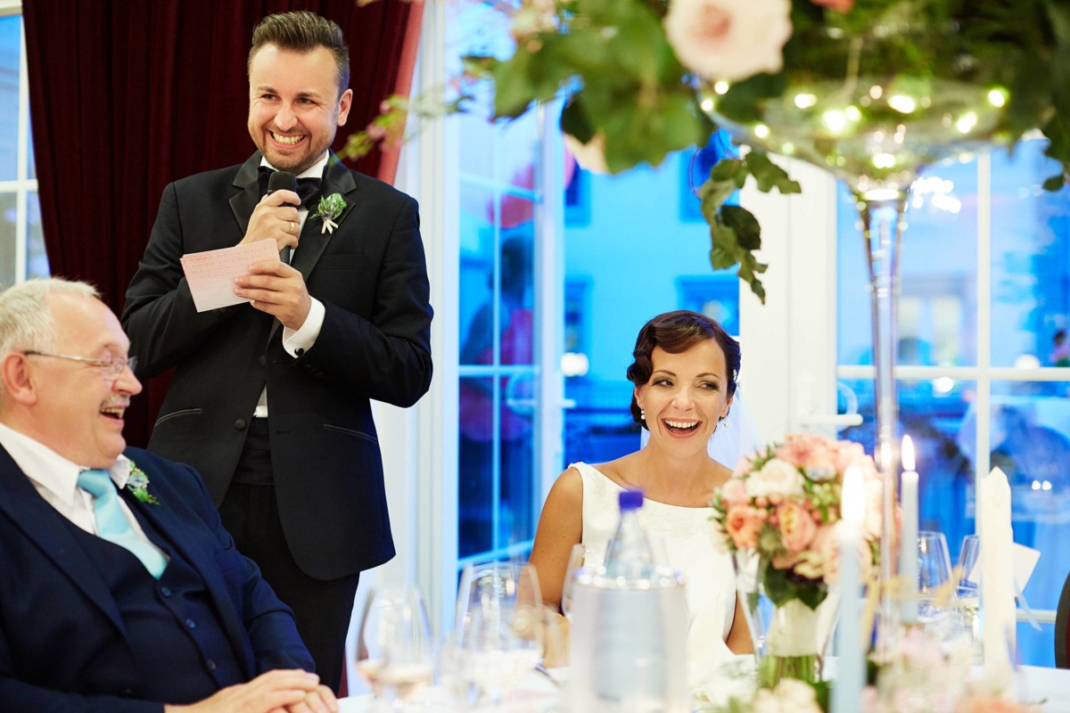 Hochzeit in einem Wellnesshotel: Bräutigam hält eine Rede, die Braut lacht laut