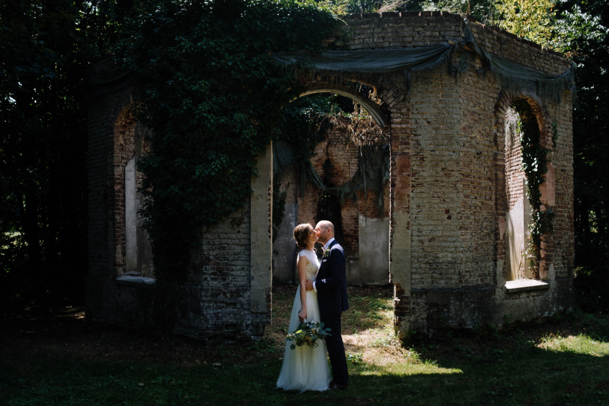 Vintage-Hochzeit: Brautpaar steht vor einer kleinen Ruine im Schlossgarten und küsst sich