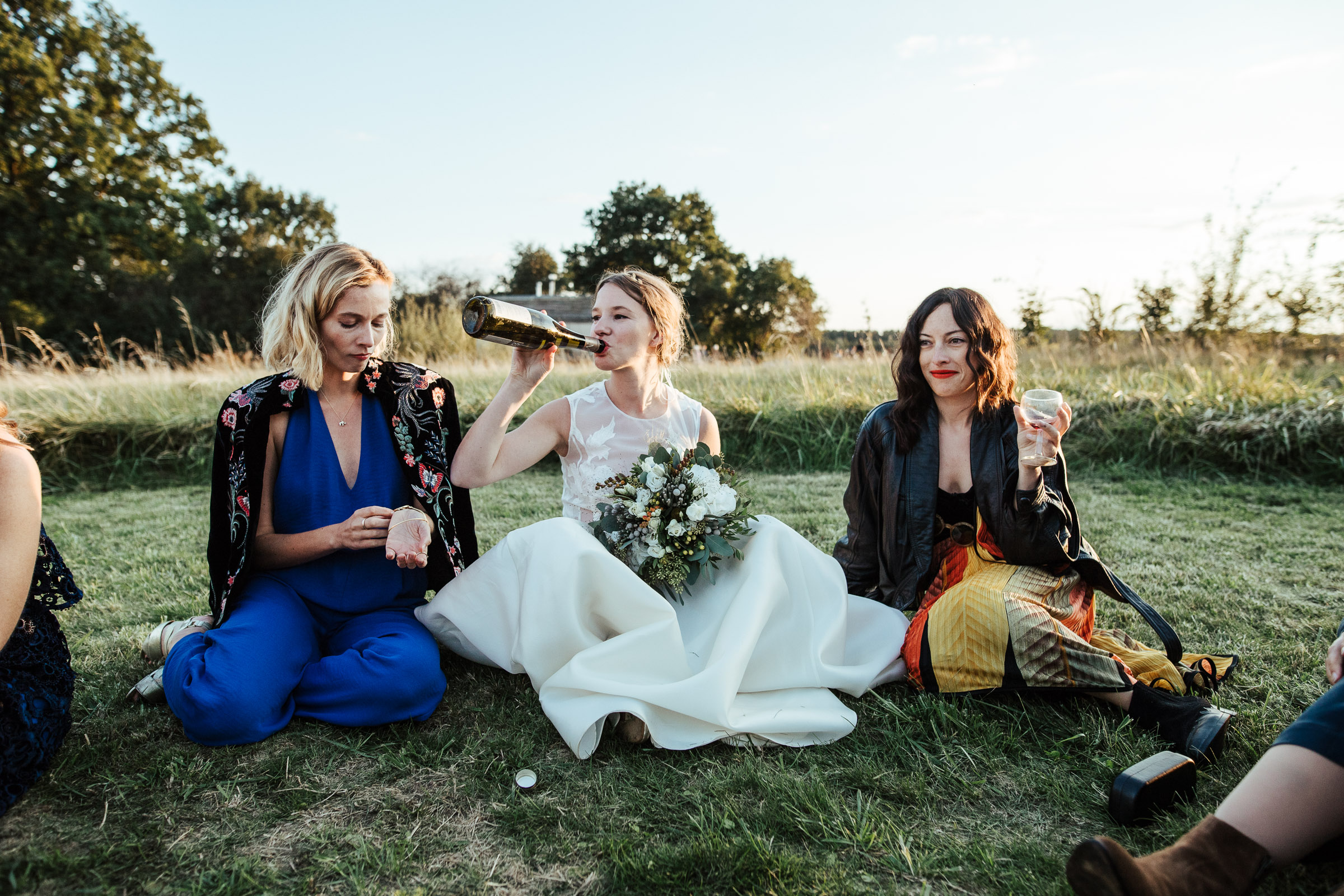 Landhochzeit: Braut trinkt direkt aus der Sektflasche mit zwei Freundinnen sitzend auf der Wiese 